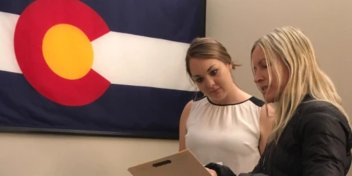 two people look at a laptop in front of a Colorado flag